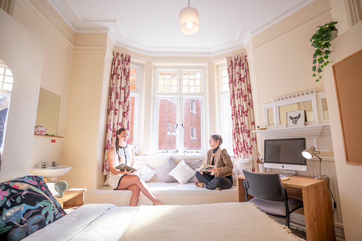 Two students chatting on a sofa ledge in a bedroom.
